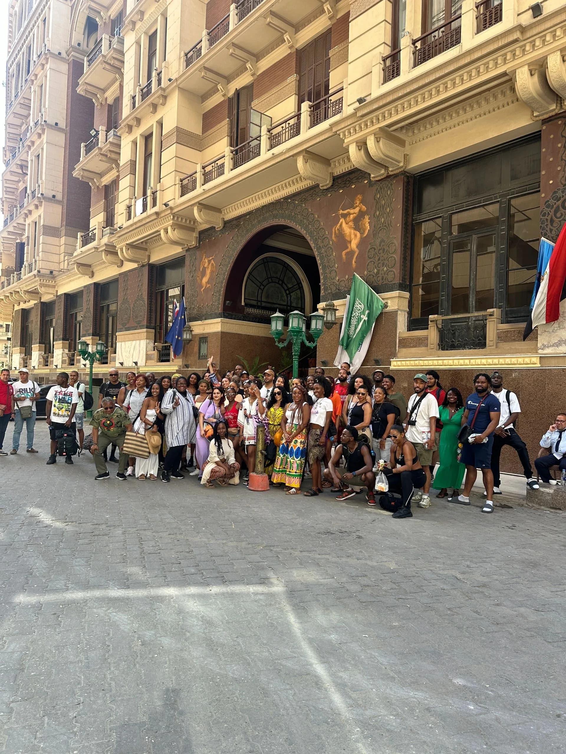 A large group of travellers gathered together on a city street in front of ornate stone buildings and flags.