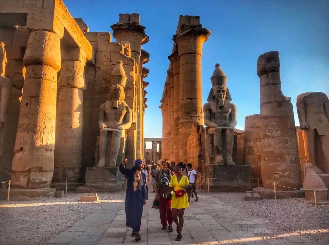 A small group of people walks among monumental carved stone columns and seated statues in warm late-day light.