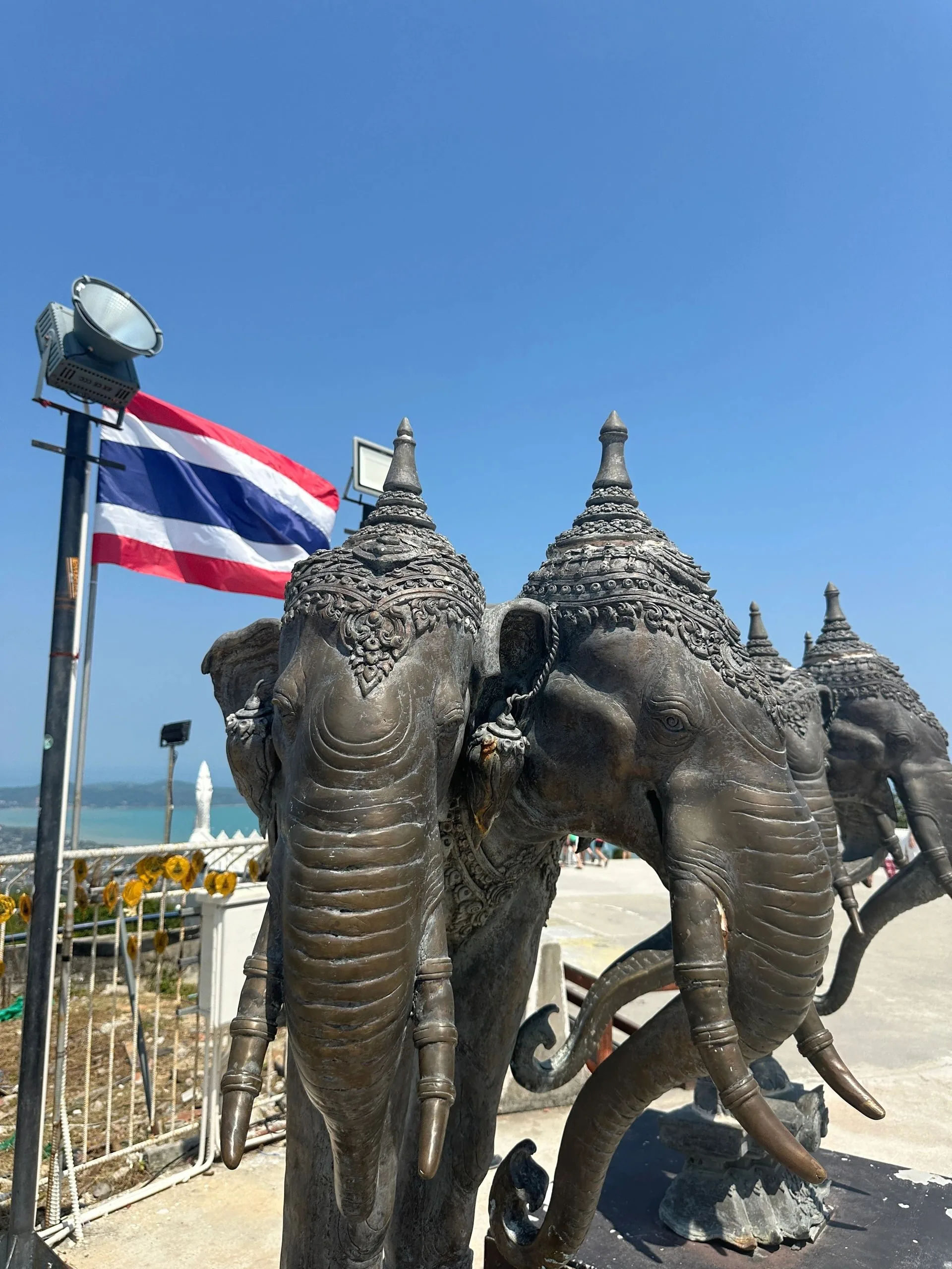 Bronze sculptures of multi-headed elephants beside a Thai flag under a clear blue sky.
